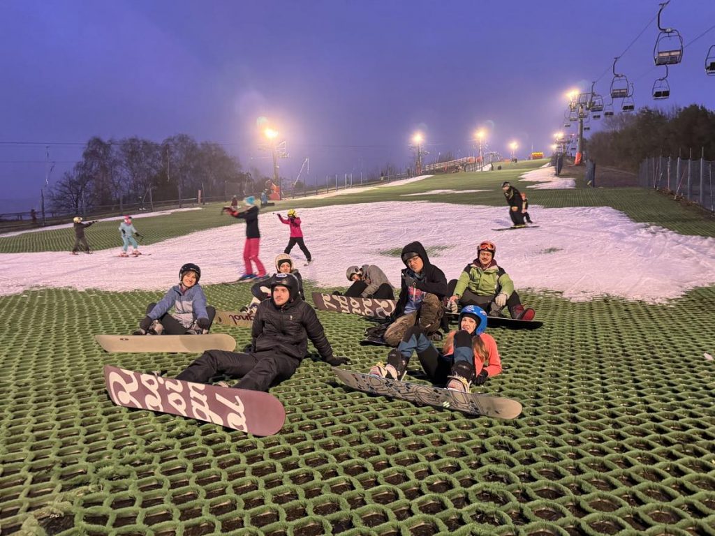 Zdjęcie przedstawia grupę młodych ludzi zjeżdżających na snowboard na sztucznej trasie