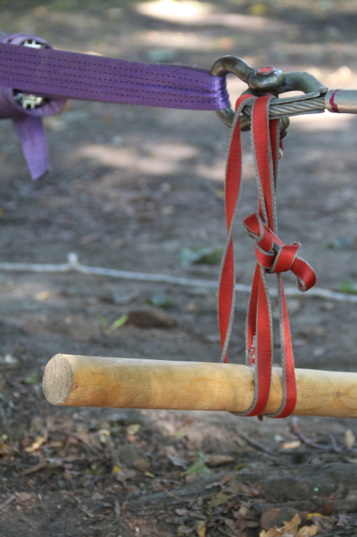 Zdjęcie przedstawia drewniany kij zawieszony przy pomocy lin w parku linowym