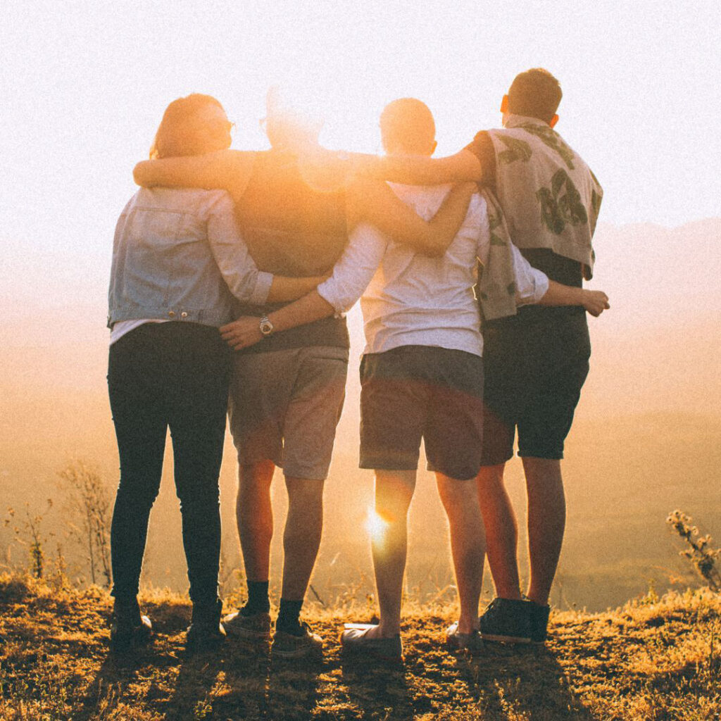 Obrazek przedstawia grupę czworga przyjaciół stojących tyłem i obejmujących się. Przyjaciele wpatrują się w zachodzące słońce.