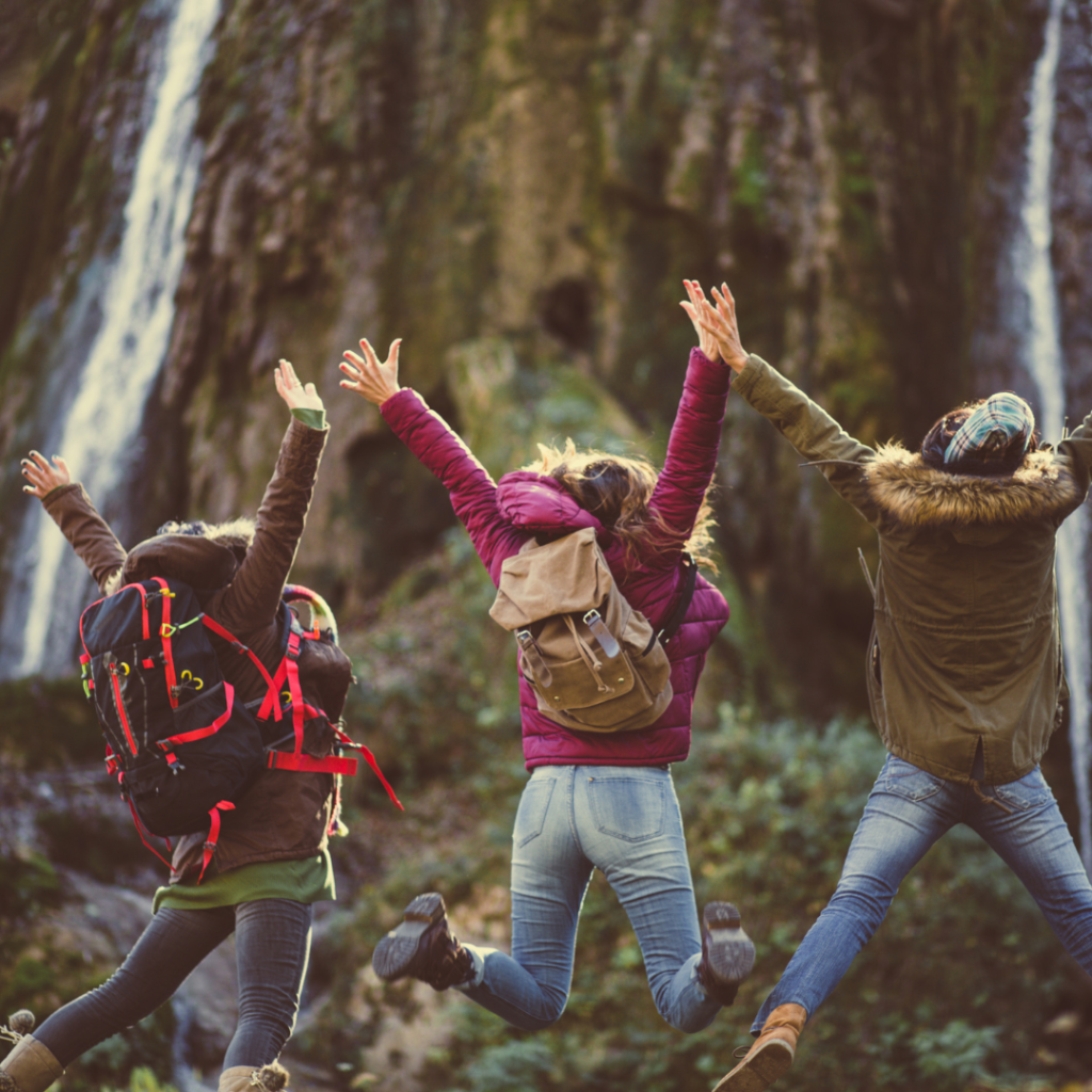 Zdjęcie przedstawia grupę przyjaciół na wyprawie w lesie, którzy trzymają się za ręce i skaczą do góry