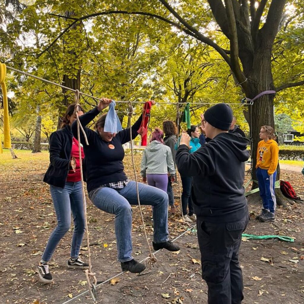 Grafika przedstawia grupę uczniów szkolnych wykonujących ćwiczenia w parku linowym pod nadzorem indstruktora