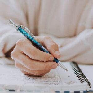 Zdjęcie przedstawia rękę trzymającą niebieski długopis nad zeszytem